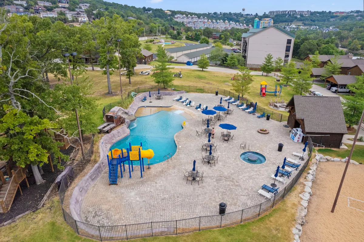 A vibrant outdoor scene displays a swimming pool, playground equipment, hot tub, lounge chairs, and a sand volleyball court, all set within a lush, green recreational area. People can be seen swimming, playing, sunbathing, and socializing, demonstrating the variety of amenities available.