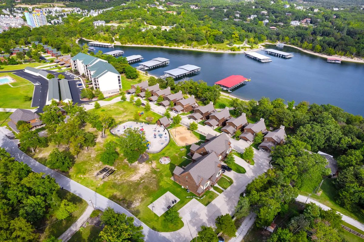 This exterior scene showcases a lush resort setting with a lake, boathouses, a playground, cabins, and expansive green spaces. People can be seen swimming, boating, picnicking, children playing, and strolling along trails. The area is vibrant with activity yet maintains a relaxed, open atmosphere.