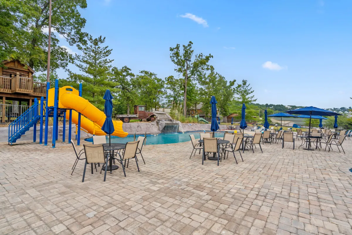 A vibrant exterior scene featuring a playground slide, a sparkling swimming pool, and an inviting outdoor seating area. Children are seen playing on the slide while families gather around tables and relax under umbrellas, emphasizing a lively and family-friendly environment.