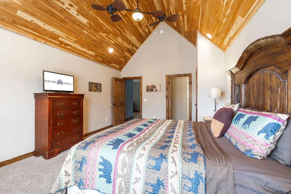 A cozy bedroom featuring a wooden ceiling, an ornate headboard, colorful bedding, and a television. The space is flooded with natural light, creating a relaxed and inviting atmosphere—ideal for restful sleep or unwinding with a book or TV.