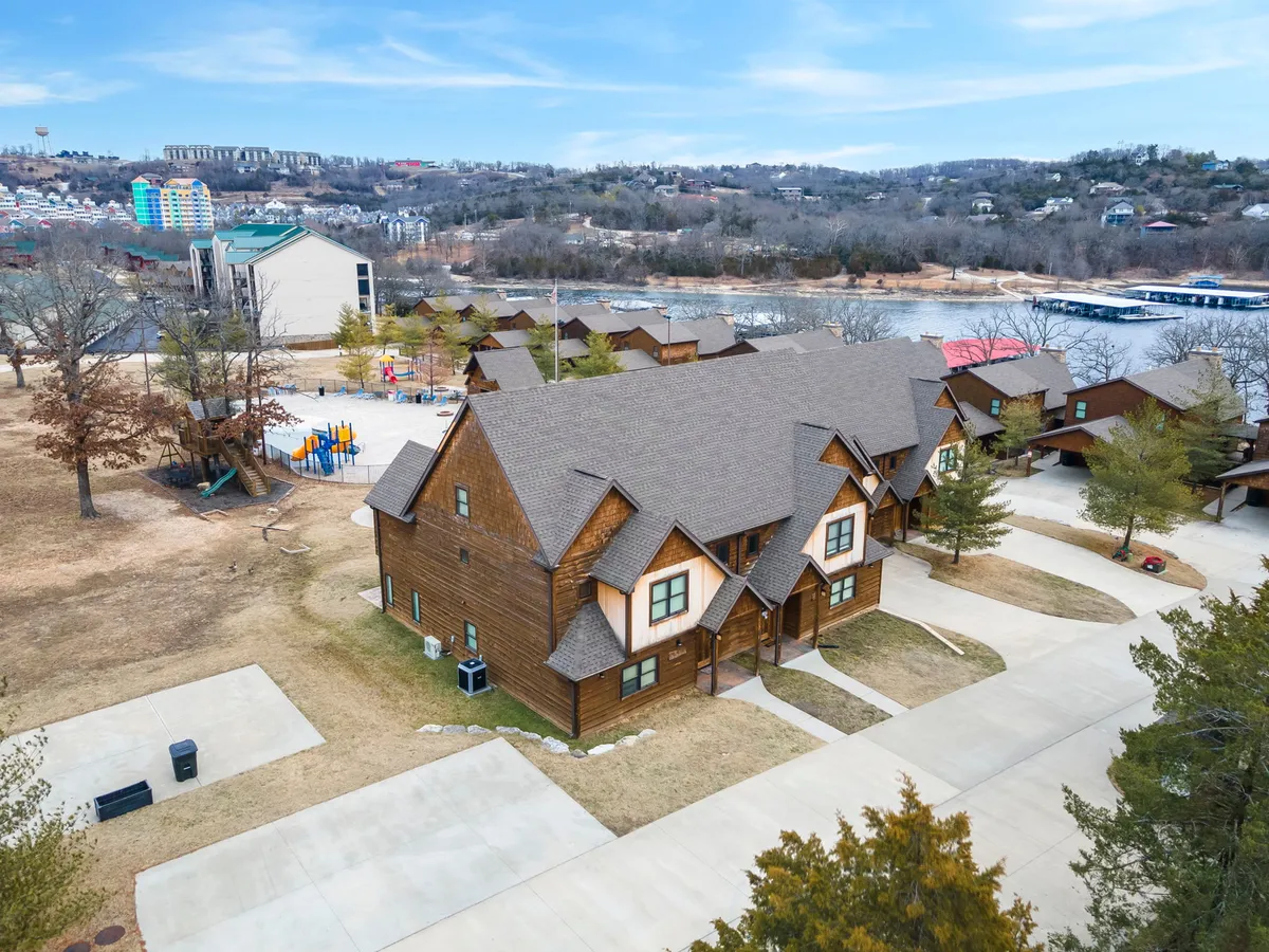 A wide exterior shot featuring rustic cabin-style buildings, a playground area bustling with activity, and a serene lake framed by trees in the background. Families gather and children play, underscoring a sense of community and outdoor enjoyment.