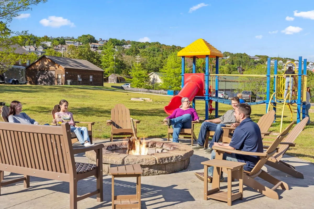 An exterior view featuring a cozy fire pit area, a nearby playground, comfortable seating, and a group of people gathered around the fire. In the background, children play on the slide while adults enjoy stories and snacks by the fire, highlighting a communal, family-friendly atmosphere.