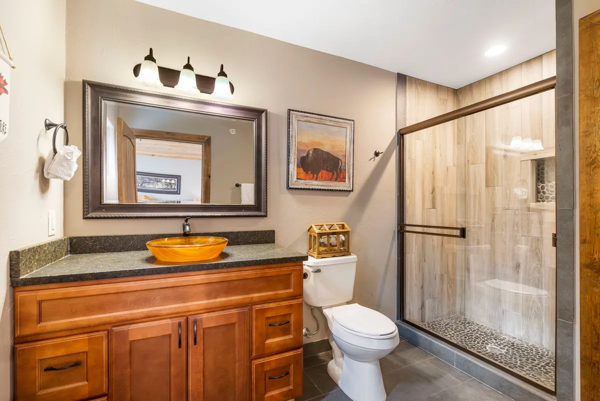 A serene, stylish bathroom with a wooden vanity, orange bowl sink, framed mirror, and large walk-in shower with glass door. The atmosphere is calm and spa-like—a perfect retreat after a long day.
