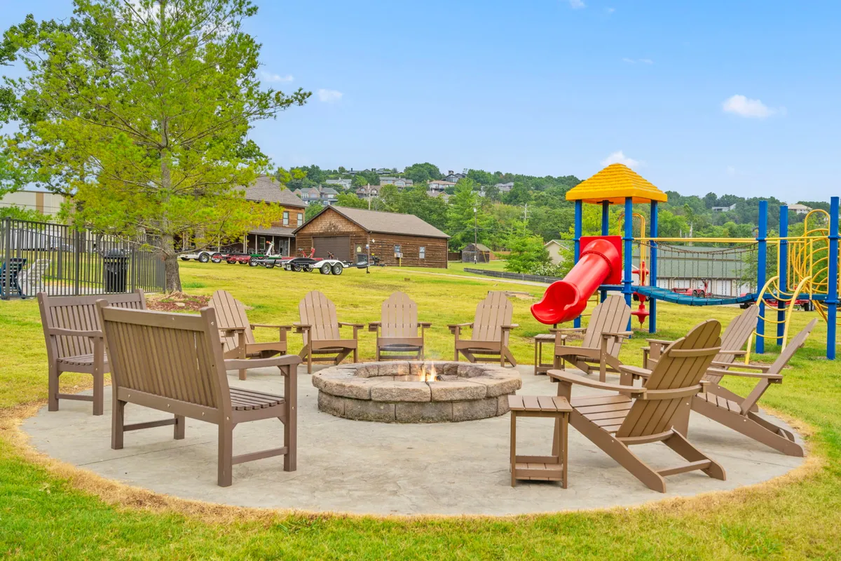 A communal fire pit is surrounded by Adirondack chairs and a playground slide sits nearby on a green lawn. The image shows people gathering around the fire and children playing, with the backdrop of an open, friendly outdoor space.