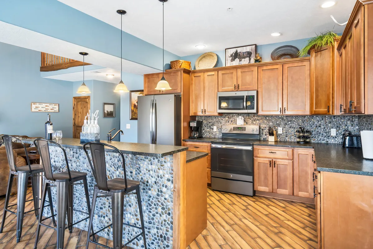 A modern kitchen featuring a large island with bar seating, stainless steel appliances, stone backsplash, and warm wood cabinetry. The setup invites culinary creativity and group meal preparation.