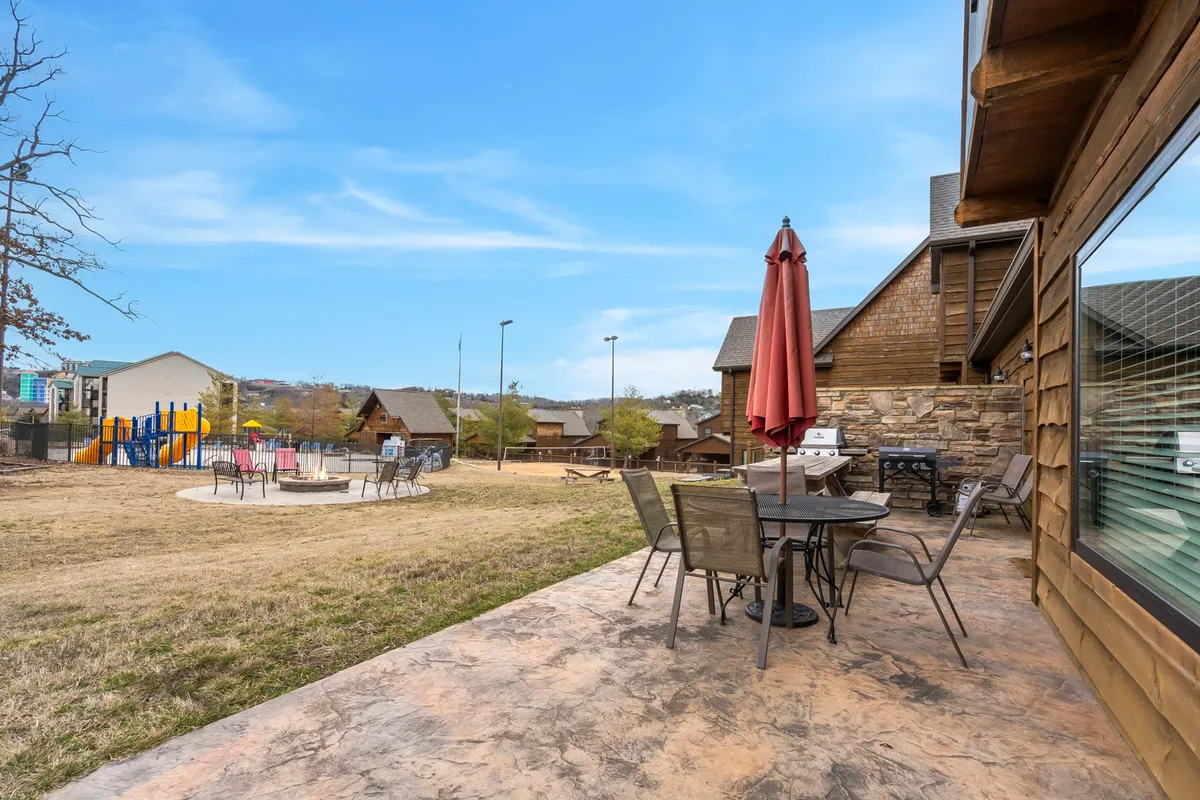 A cheerful outdoor patio with comfortable furniture and a red umbrella, overlooking a playground and surrounded by cozy cabins. The area is clearly suited for both human and canine activities, suggesting ample space for pets to play safely and guests to enjoy the outdoors.