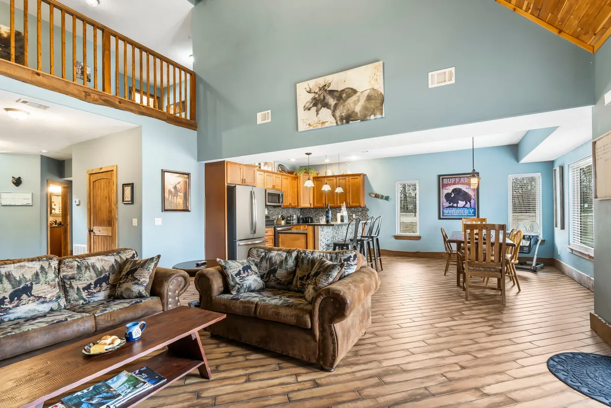 A bright and welcoming open-plan living area featuring a sofa with a wildlife print, a wood-finished kitchen, a dining area, and expansive windows that fill the space with natural light. The arrangement encourages socializing, relaxation, and connection for both family and pets.