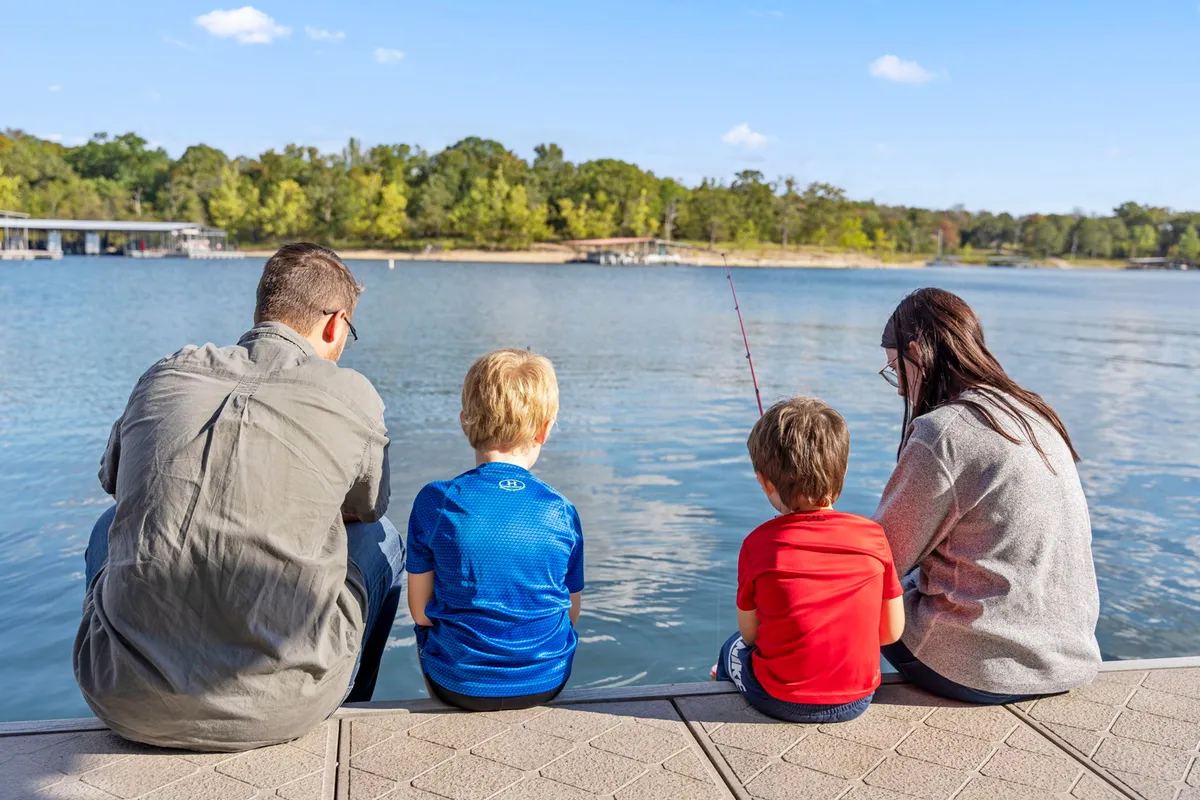 A family stands together at the edge of the lake, with children and adults engaged in fishing. The water is calm, and the expressions show joy and bonding, illustrating the experience of creating memories by the lake.