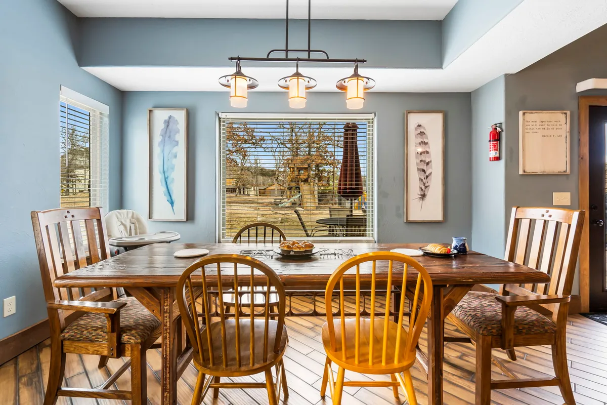 The image showcases a spacious, warmly lit dining area with a large table set beneath elegant light fixtures. Sunlight pours in through expansive windows, offering a glimpse of the scenic outdoors, while tasteful artwork and inviting décor create a cozy, welcoming atmosphere perfect for gathering.