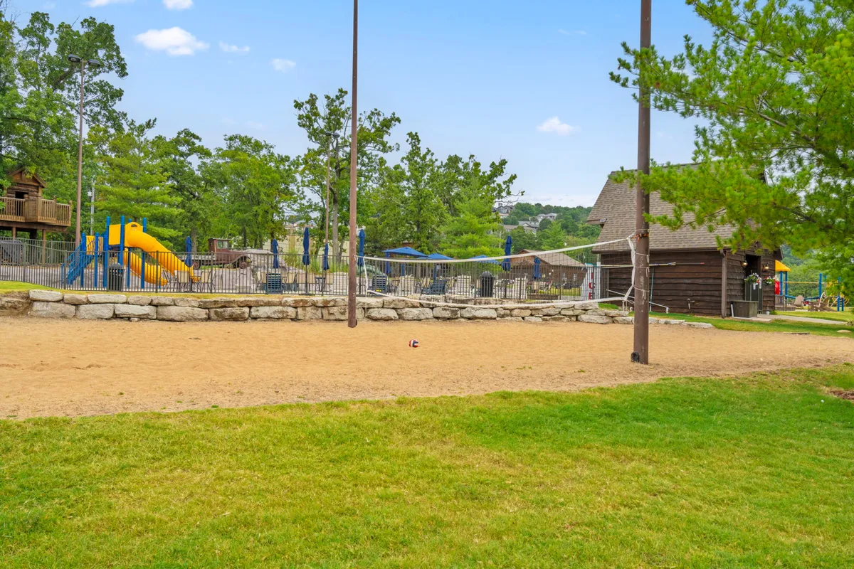 A cheerful outdoor setting featuring children playing on the playground, families picnicking, and a sand volleyball court set among mature trees. The scene highlights year-round amenities with a focus on community, activity, and family-friendly fun.