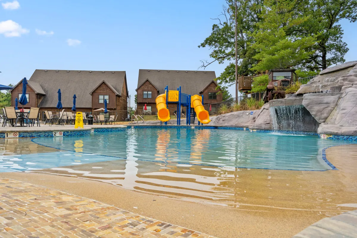 An inviting outdoor swimming pool features a water slide and a cascading waterfall. Surrounding the pool are lounge chairs and shaded seating areas, with families and children enjoying the water and relaxing nearby. The playful atmosphere and lush landscaping evoke the feeling of a resort getaway.